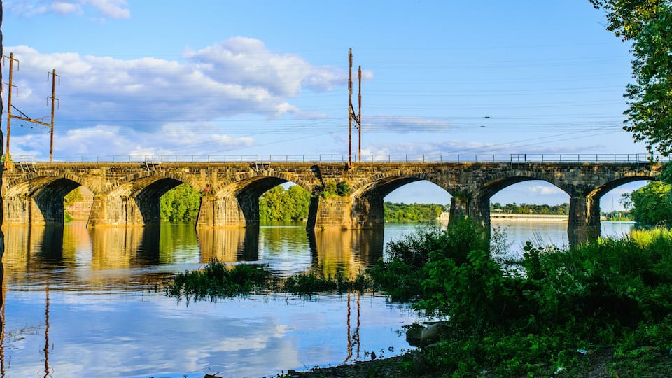 A stone bridge with multiple arches spanning a river, surrounded by greenery and power lines.