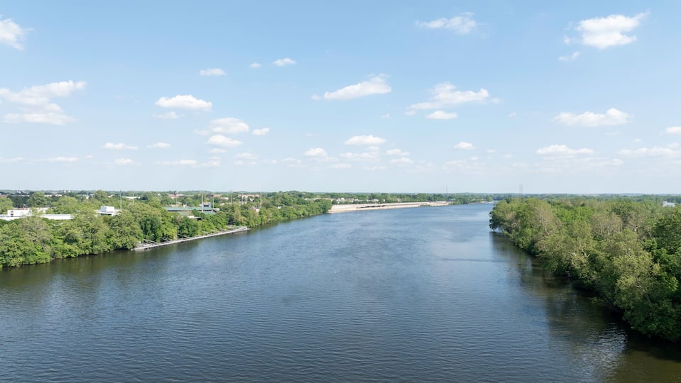 A river flanked by dense green foliage and a clear blue sky with scattered clouds.
