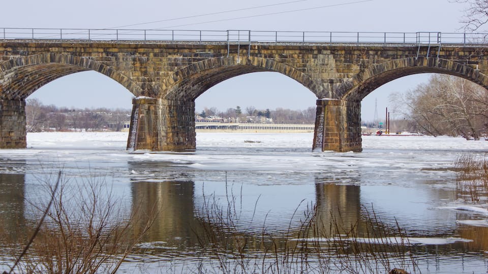 A stone bridge over a frozen river with bare trees and a distant building.