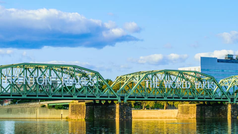 A green steel bridge with the words "Trenton Bridge" and "The World Takes" on it, spanning over a river with a modern building in the background.