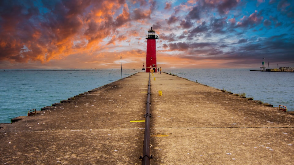 A red lighthouse on a pier with a sunset sky.
