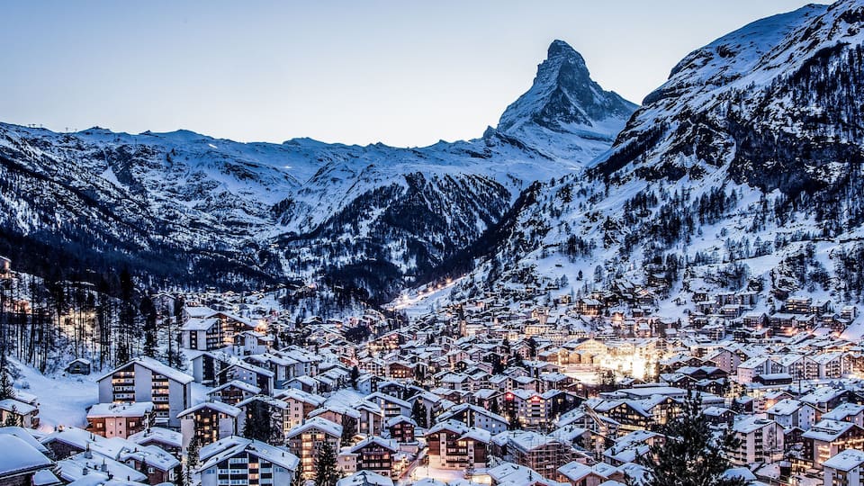 A snow-covered mountain town with illuminated buildings and a prominent peak in the background.