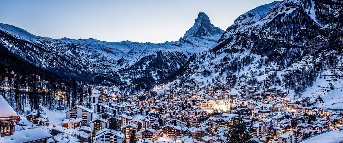 A snow-covered mountain town with illuminated buildings and a prominent peak in the background.