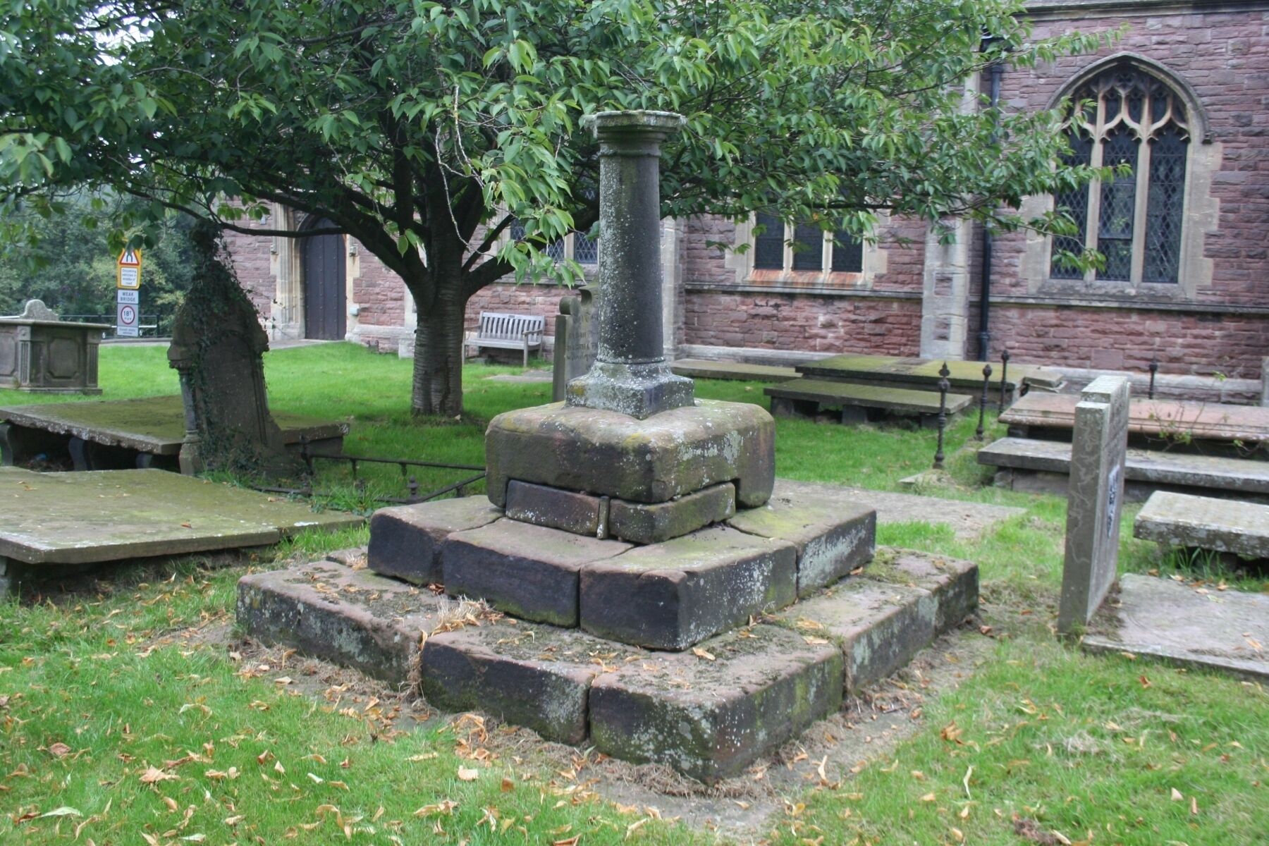 A stone monument with a central pillar, surrounded by grass and trees, in a cemetery with stone benches and a brick building in the background.