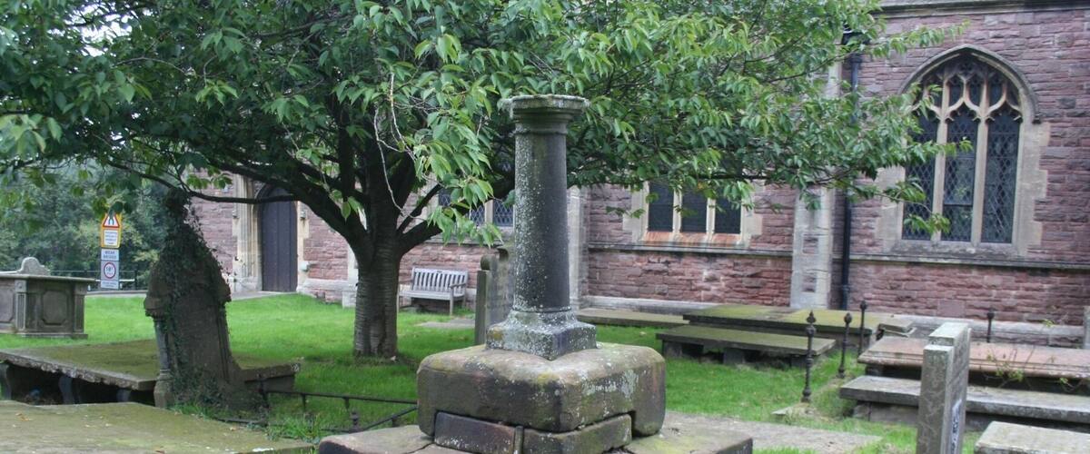 A stone monument with a central pillar, surrounded by grass and trees, in a cemetery with stone benches and a brick building in the background.