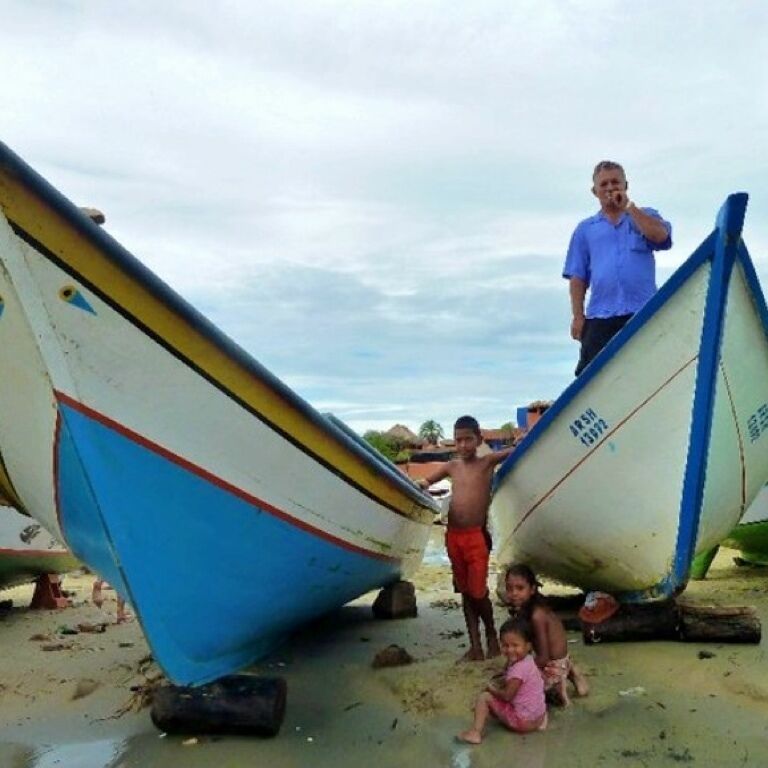 A beach with several boats, a man in a blue shirt, and two children.