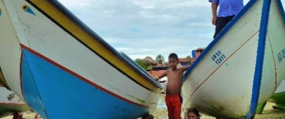 A beach with several boats, a man in a blue shirt, and two children.
