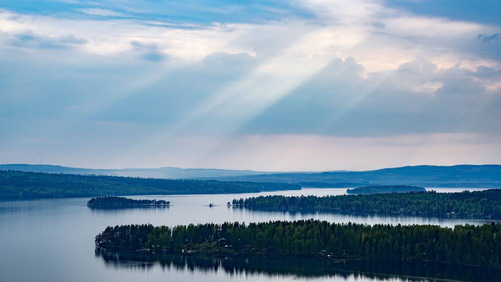 A calm lake surrounded by dense forests and distant hills under a cloudy sky.