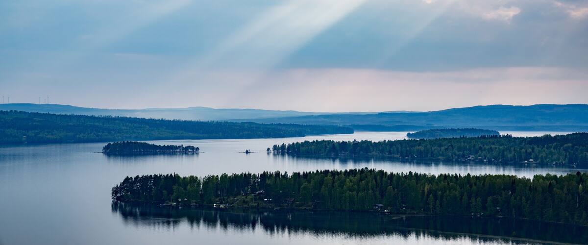 A calm lake surrounded by dense forests and distant hills under a cloudy sky.