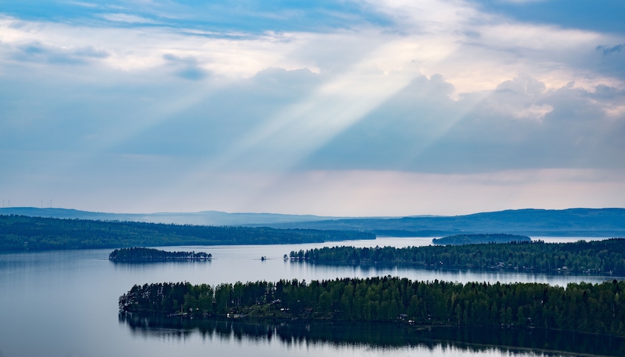 A calm lake surrounded by dense forests and distant hills under a cloudy sky.