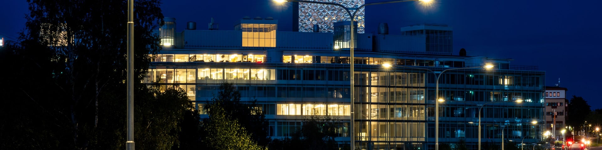 Um edifício moderno com fachada geométrica marcante, cercado por árvores e uma rua bem iluminada à noite.