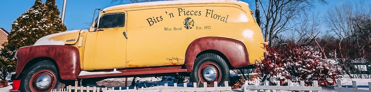 A vintage yellow truck with 'Bits 'n Pieces Floral' branding parked in a snowy setting with a flagpole and trees in the background.