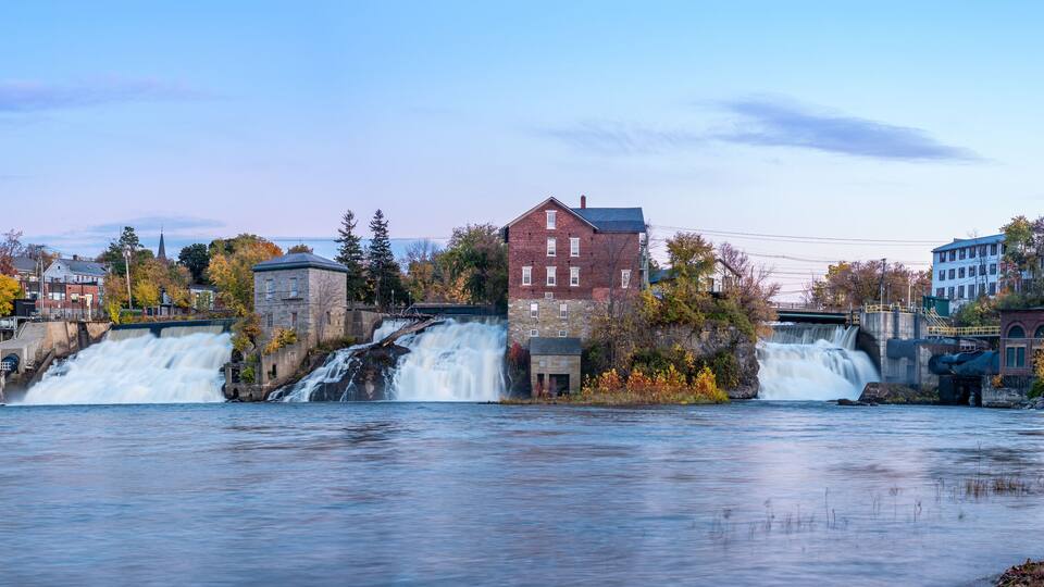 A river with multiple waterfalls, surrounded by autumn-colored trees and buildings.