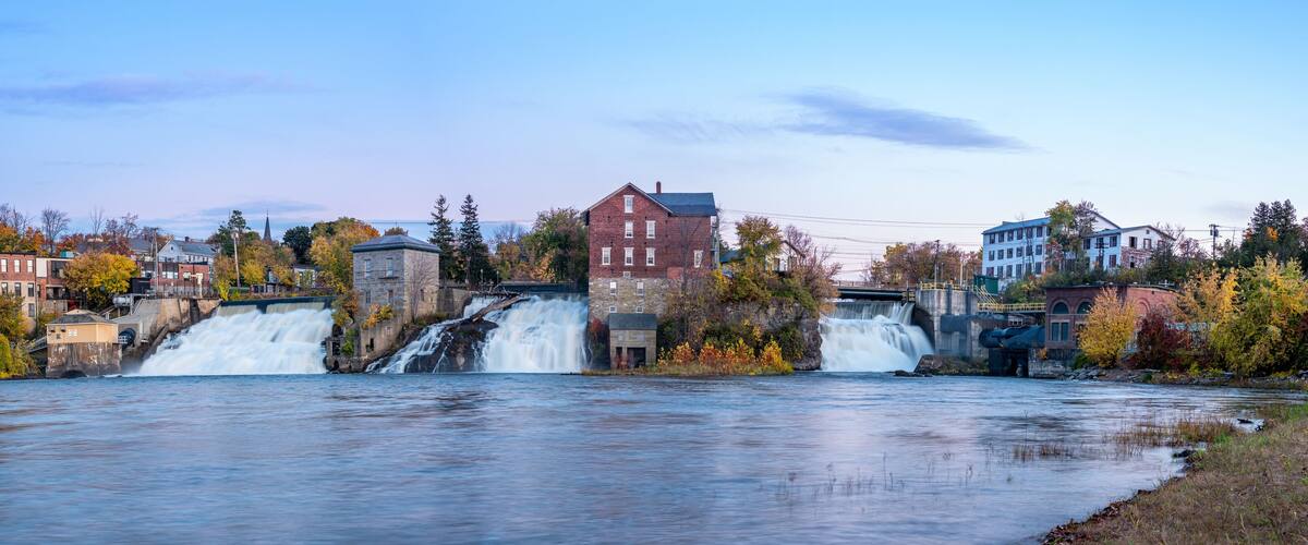 A river with multiple waterfalls, surrounded by autumn-colored trees and buildings.