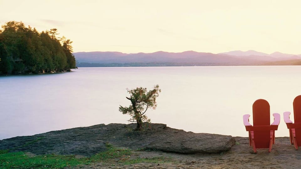 A lake with mountains in the background and two red Adirondack chairs on a rocky shore.