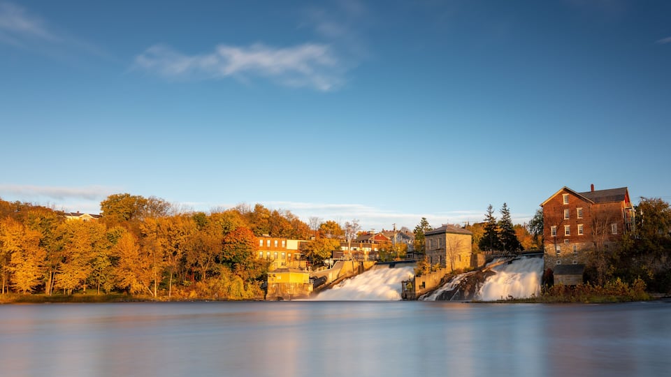 A river with a waterfall, autumn trees, and buildings along the riverbank.