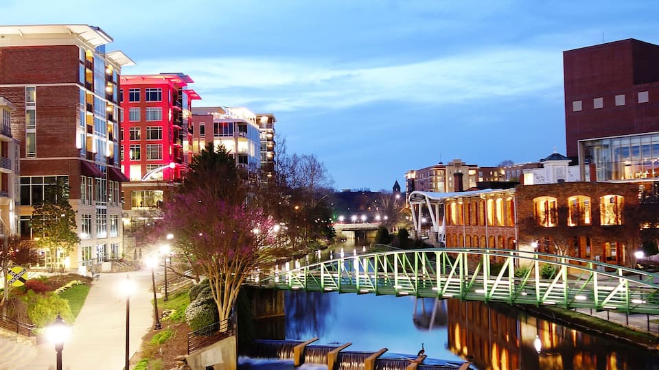 A riverside walkway with a bridge, buildings, and a waterfall.