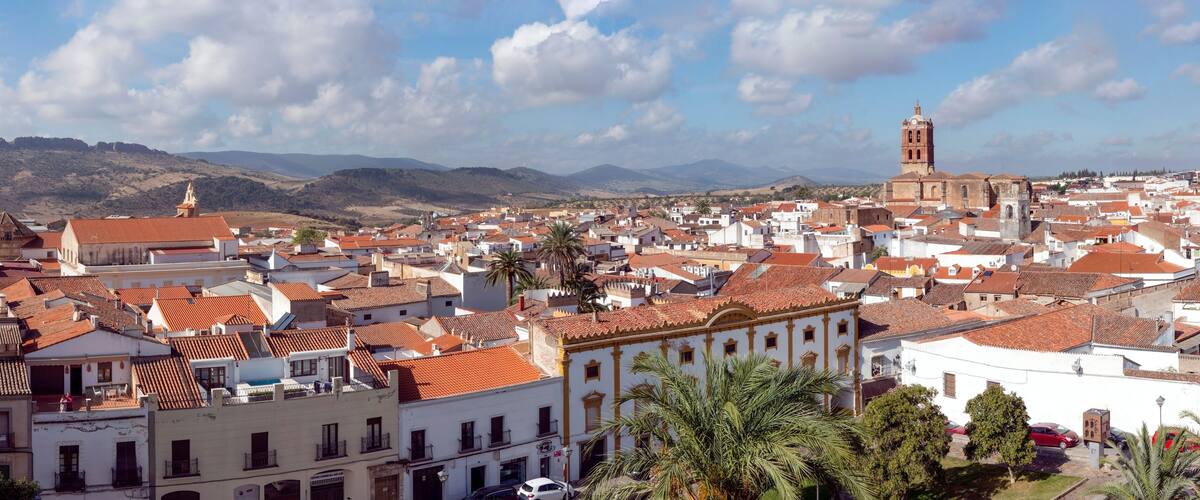 A town with red-tiled roofs, a prominent church with a bell tower, and mountains in the background.