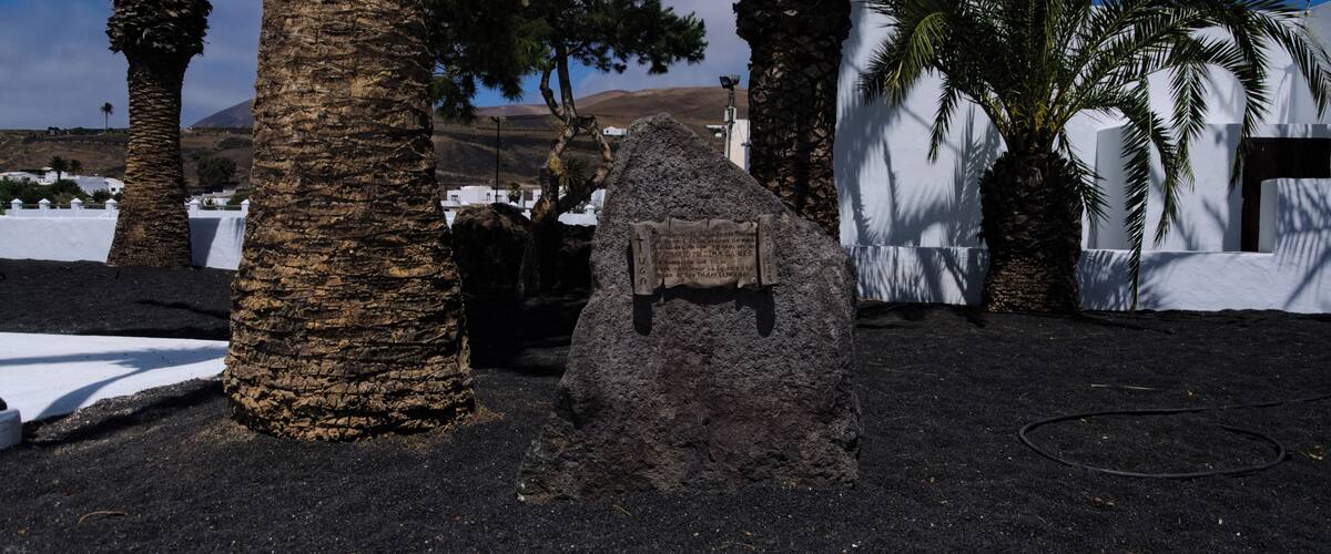 A rocky formation with a plaque, palm trees, and white buildings in the background.
