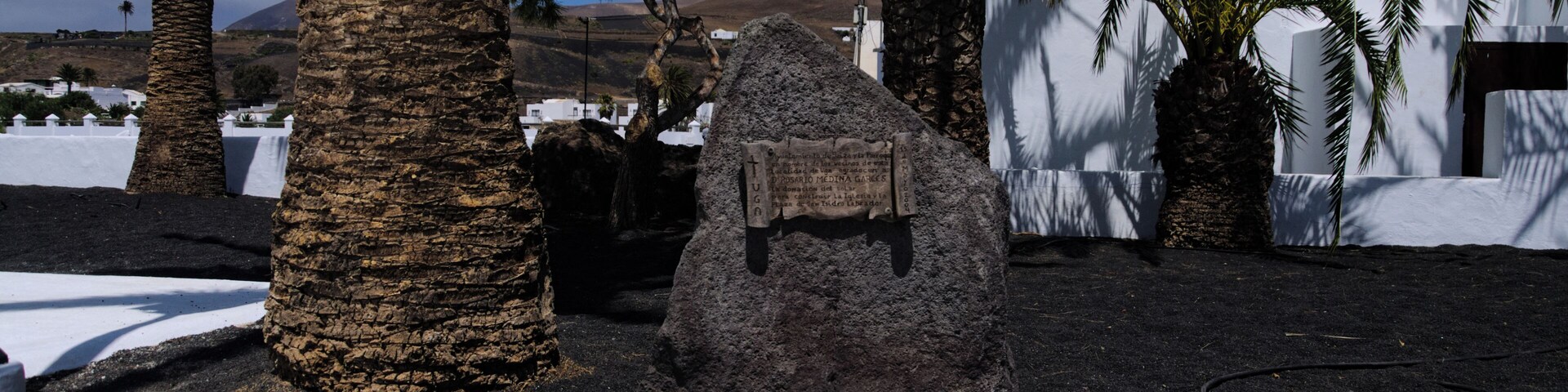 A rocky formation with a plaque, palm trees, and white buildings in the background.