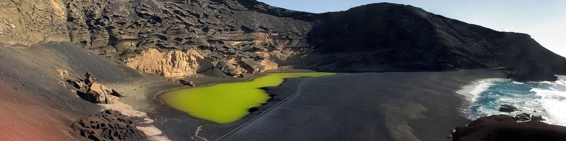 A coastal landscape with a black sand beach, green pools, and rocky cliffs.