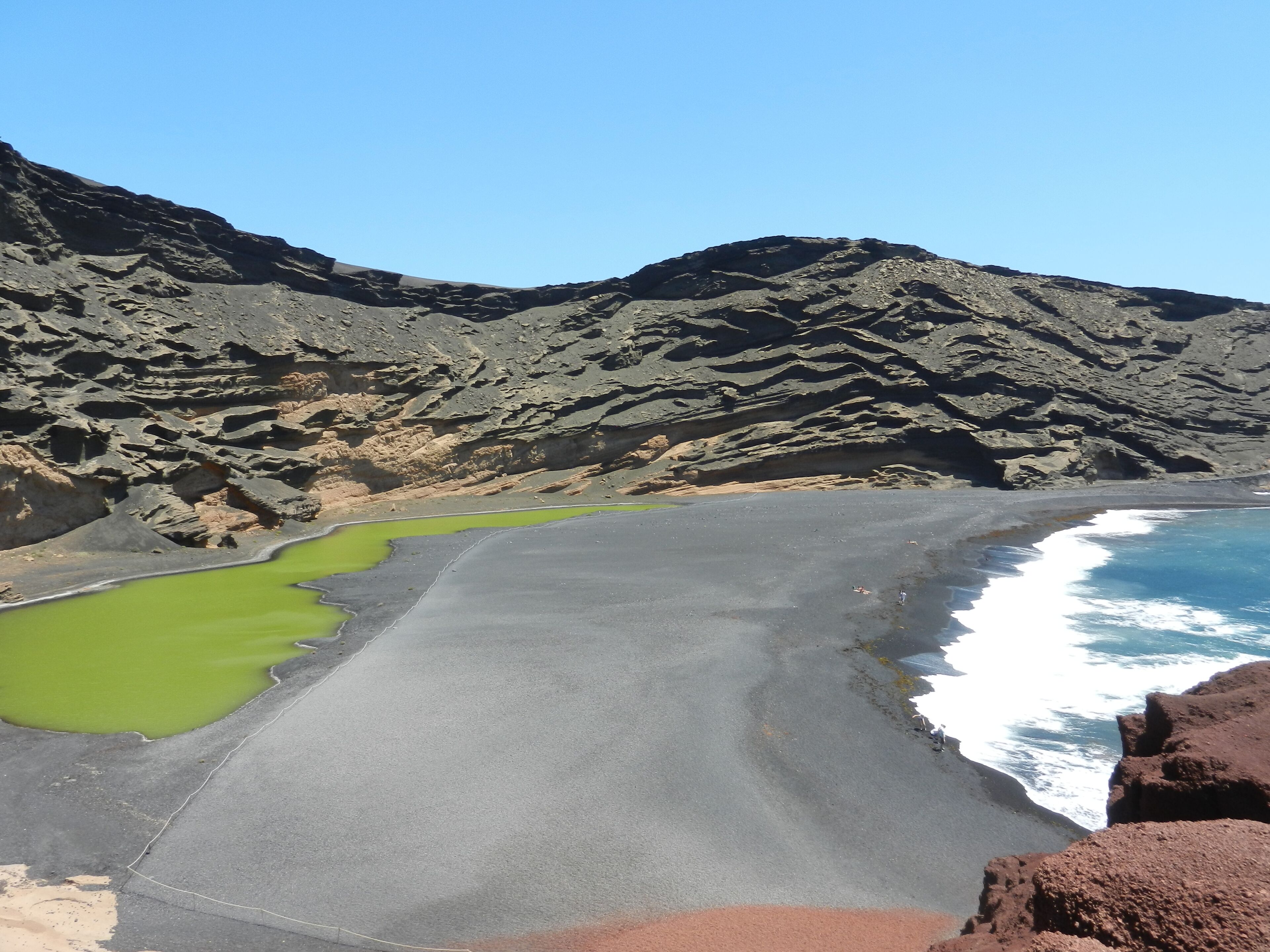 Una playa con albercas verdes, orilla de arena negra y acantilados rocosos.