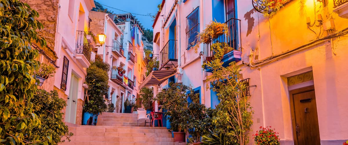 A narrow street with steps leading up to a building, surrounded by colorful buildings and potted plants.
