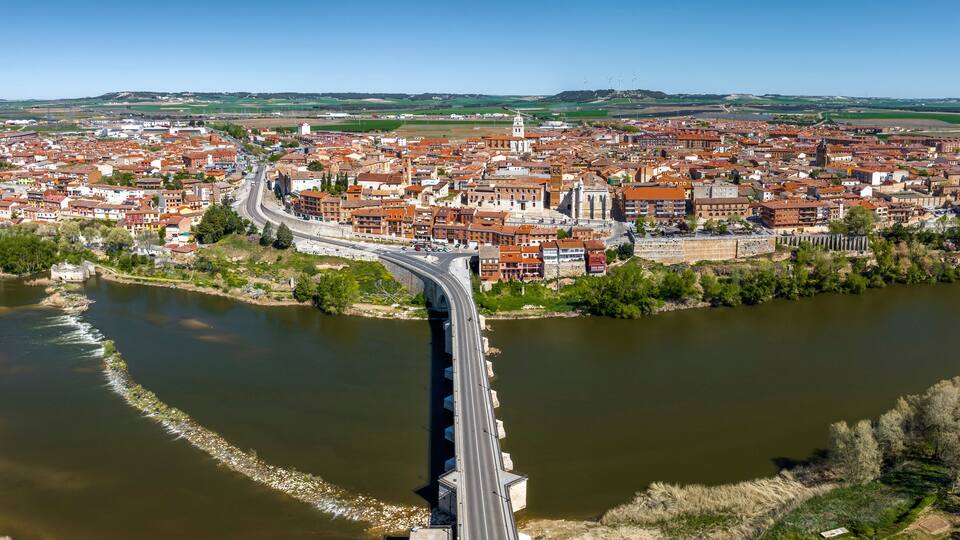 A river with a bridge, a town with red-roofed buildings, and green fields in the background.