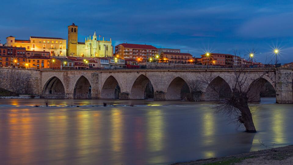 A stone bridge over a calm river at twilight with illuminated buildings and a prominent tower in the background.