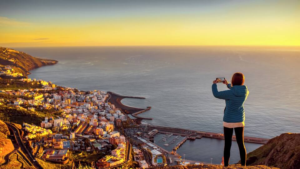 Young female traveler in blue jacket photographing Santa Cruz city standing on the top of the mountain on La Palma island in the morning