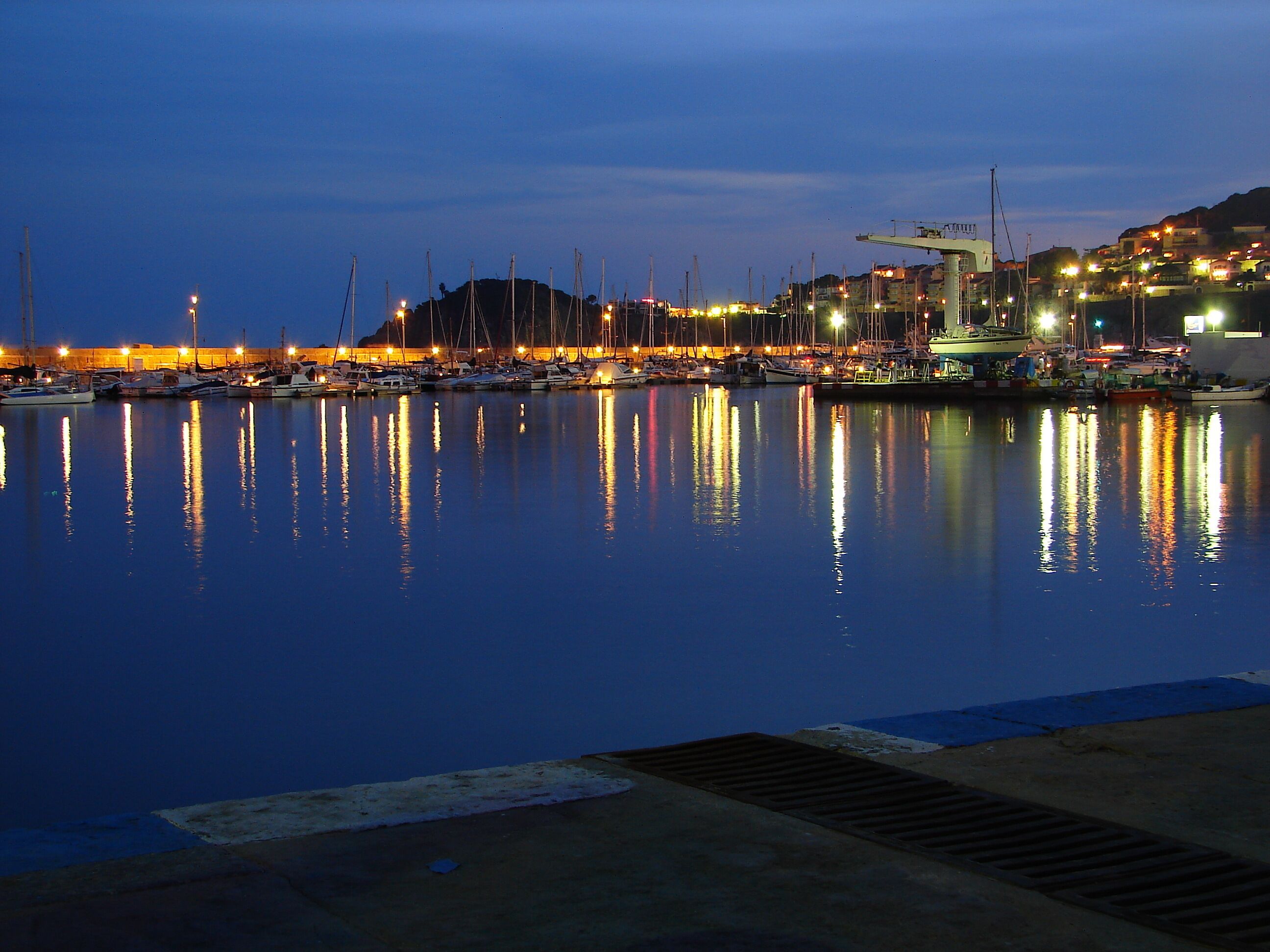 Un puerto deportivo al atardecer con barcos amarrados y luces de la ciudad reflejándose en el agua.