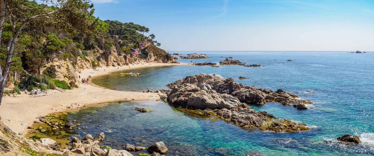 A coastal landscape with clear blue waters, rocky outcrops, and a sandy beach.