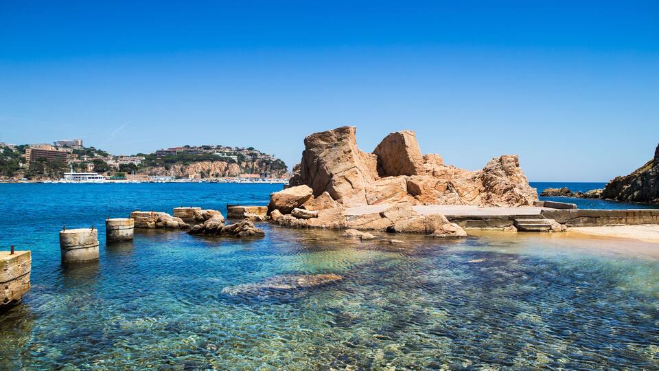 A coastal landscape with clear blue water, rocky formations, and a distant cityscape.