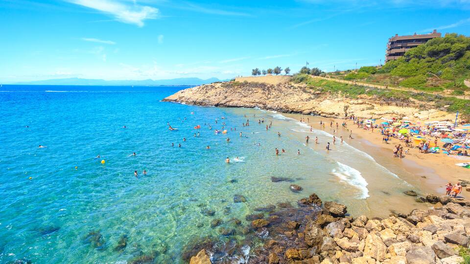 A beach with clear blue water, rocky shoreline, and a building on a hill.