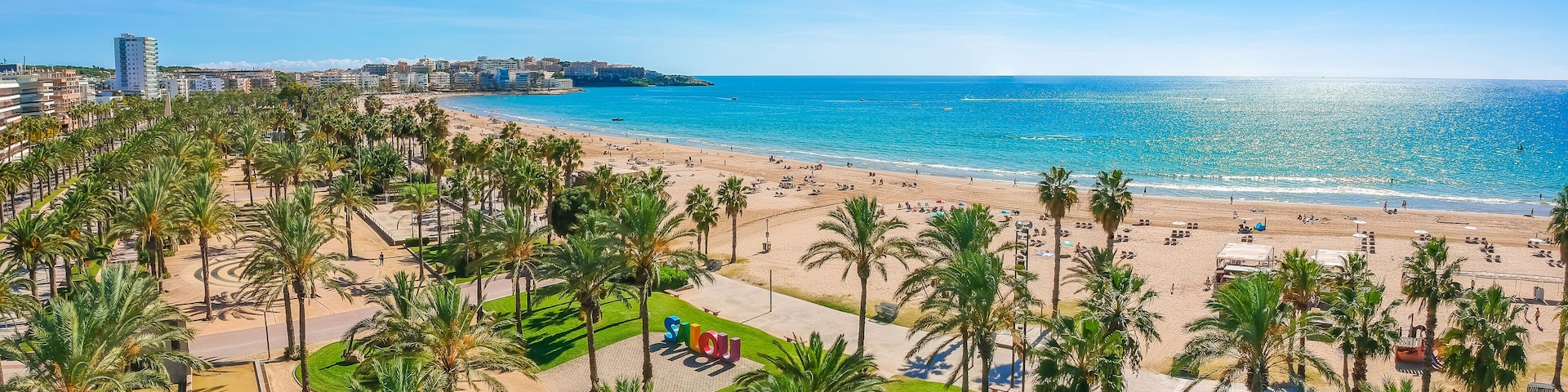 A coastal cityscape with a beach, palm trees, and a clear blue sky.