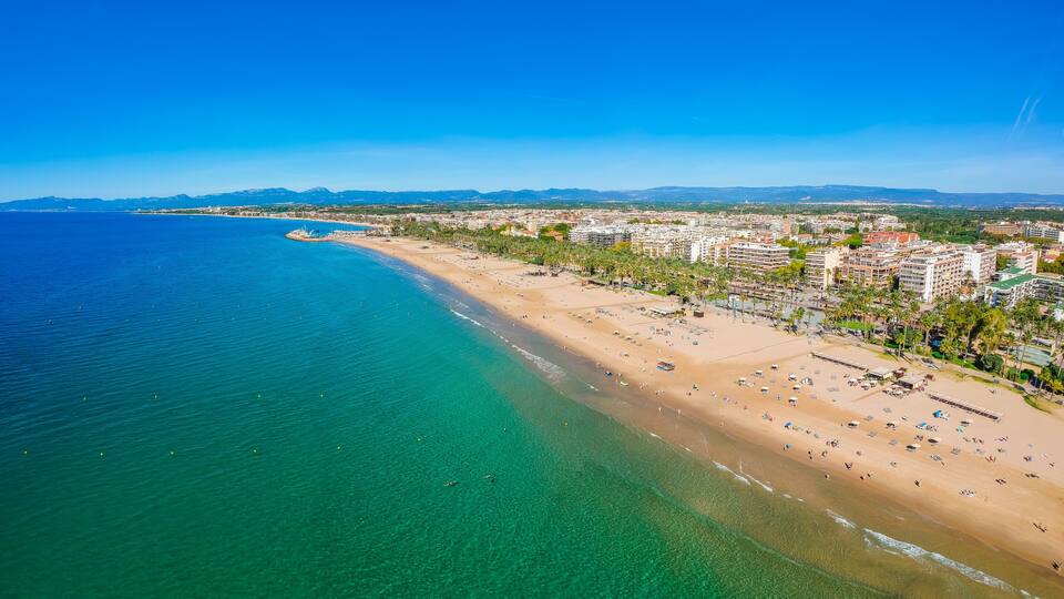 A coastal cityscape with a wide beach, clear blue waters, and distant mountains.