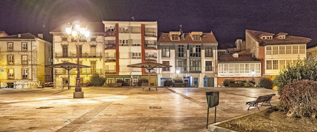 A nighttime urban square with buildings, street lamps, and benches.