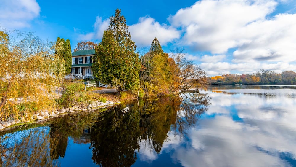 Ein Haus am Ufer eines stillen Sees, umgeben von Herbstlaub und bewölktem Himmel.