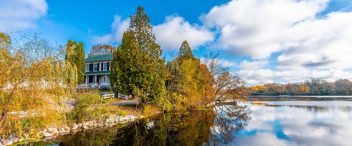 A house by a calm lake with autumn foliage and a clear sky.