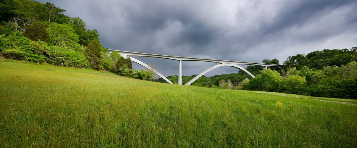 A white bridge over a grassy field with a forested hill in the background and a cloudy sky.