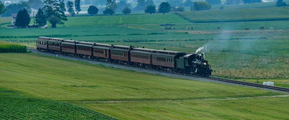 A steam train moves along the tracks surrounded by green fields and trees. The sun shines down as the train passes through a rural landscape, creating a scenic view.
