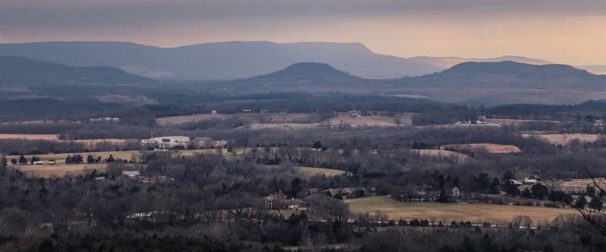 A valley with rolling hills, scattered houses, and a distant mountain range under a cloudy sky.