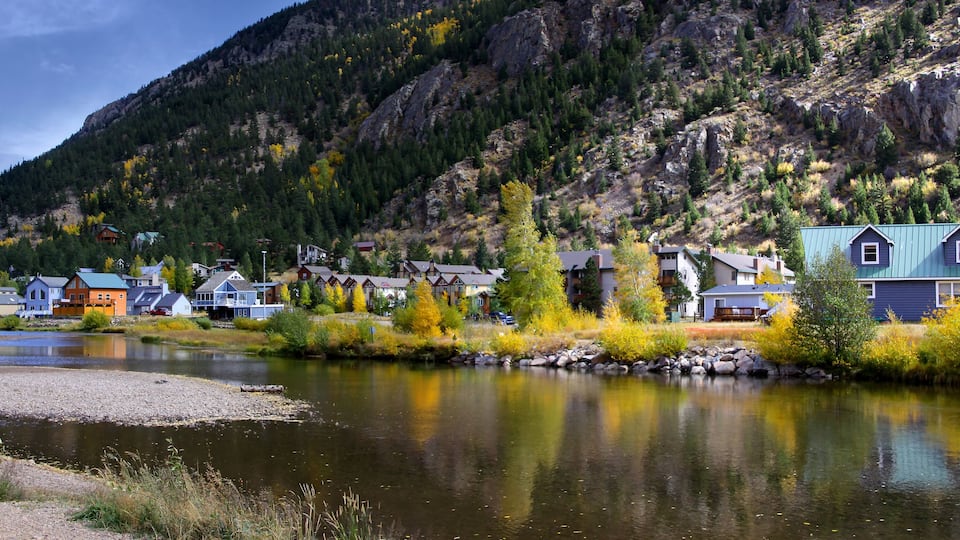 A river with houses along its banks, a mountain in the background, and autumn-colored trees.
