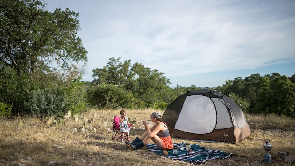 A camping scene with a tent, a child in a pink chair, and a woman on a blanket.