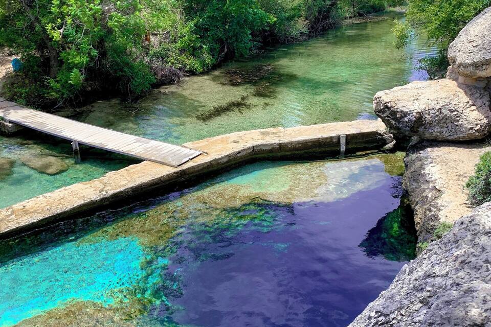 Un plan d’eau aux eaux claires, surmonté d’un pont en bois, et entouré d’une végétation luxuriante et de rochers.