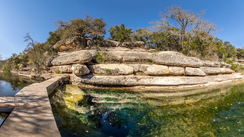 A rocky island with a clear pool of water reflecting the sky and surrounding trees.