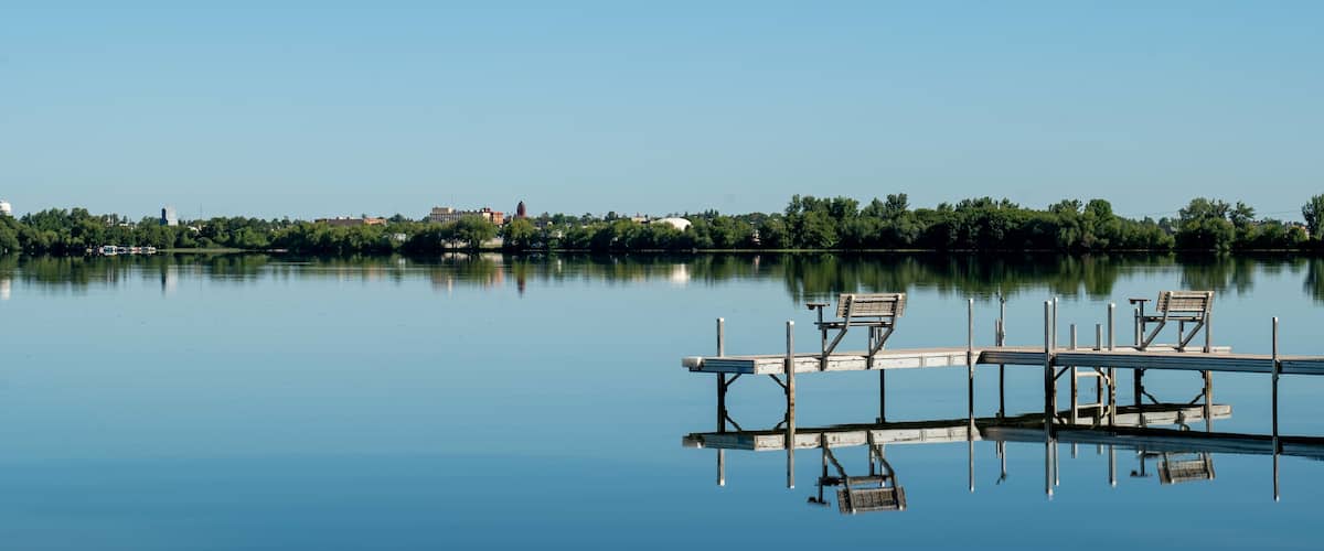 An inviting boat dock is reflected in the glassy water of Lake Irving, the first lake on the Mississippi river, with Bemidji, Minnesota, the 2018 Best Town in Minnesota, seen across the lake.