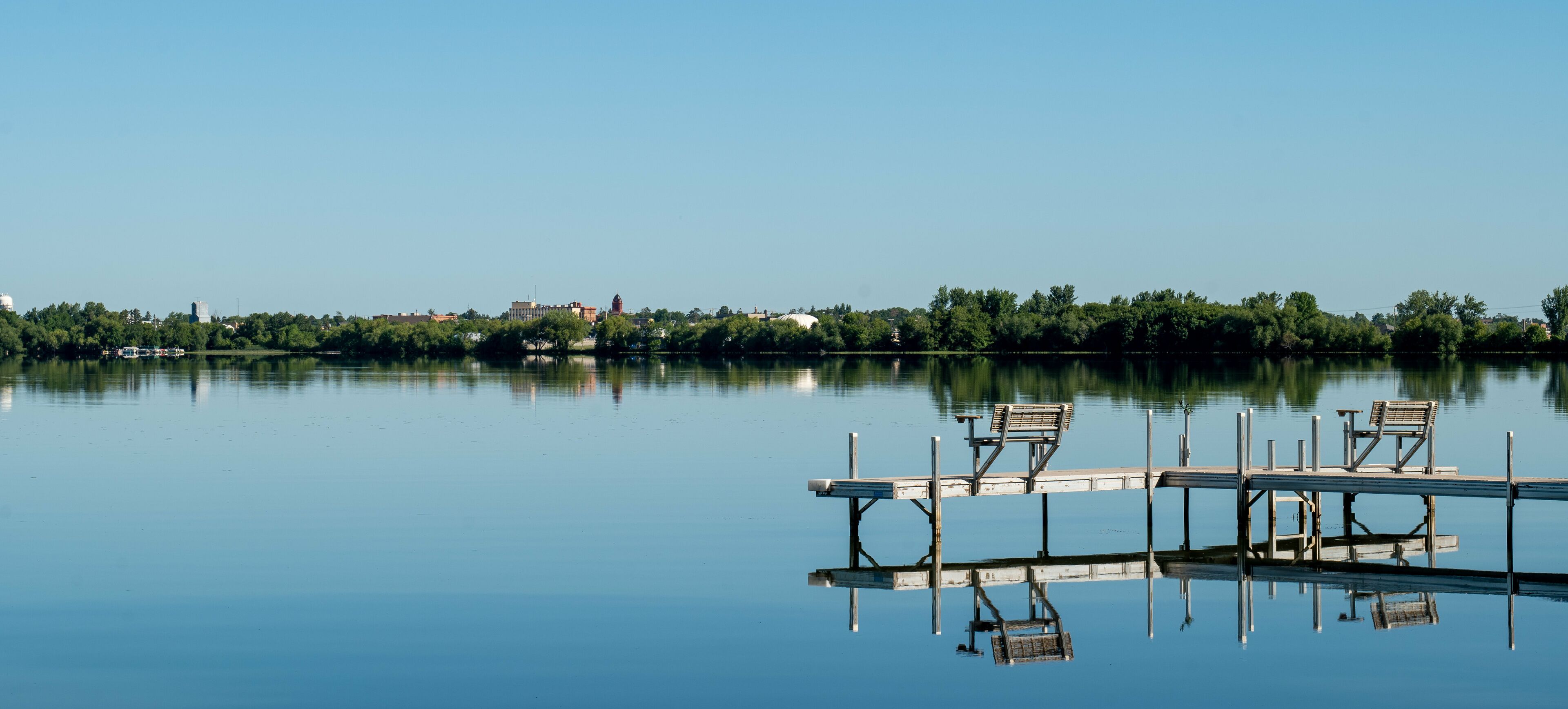 A calm lake with a wooden dock and benches, surrounded by trees and buildings in the distance.