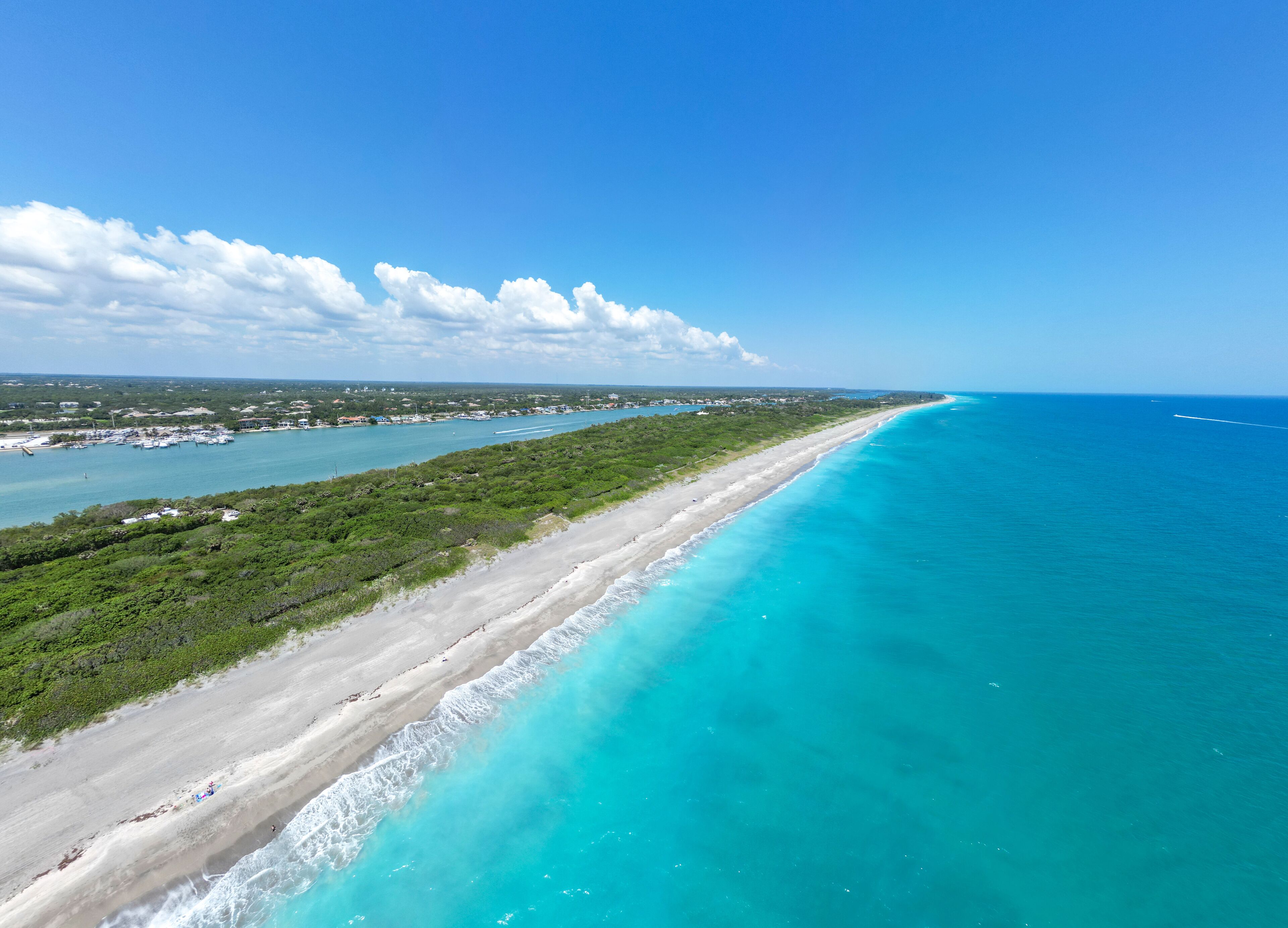 A coastal landscape with a long sandy beach, turquoise waters, and a marina with boats.