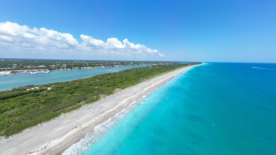 A coastal landscape with a long sandy beach, turquoise waters, and a marina with boats.